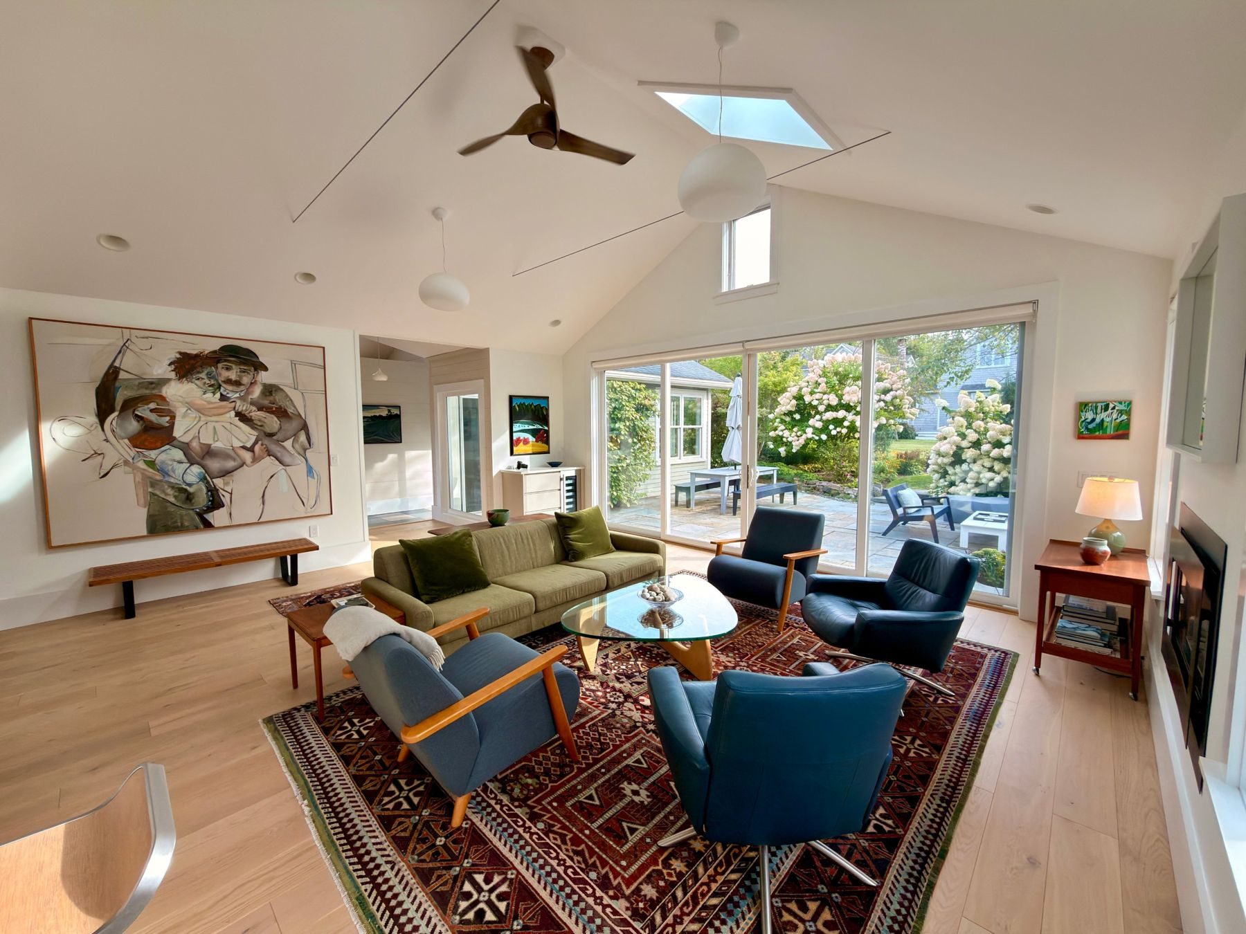 The Mansard's sunroom: vaulted ceiling and skylight, a large figurative painting, blue chairs on a vintage rug, and glass doors open to the hydrangea garden