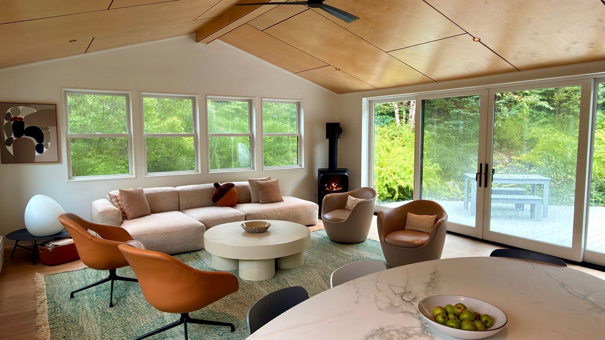 The Garage living room: a birch-paneled vaulted ceiling, linen sofa, cognac leather chairs, wood stove and glass doors to the fern-lined deck