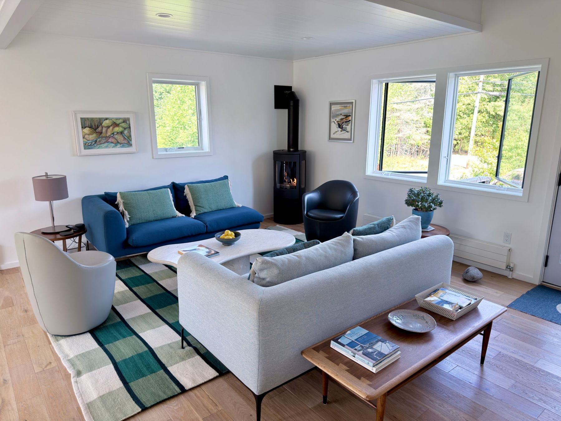 Schoolhouse living room: blue sofa, gas stove, green plaid rug and windows to the trees